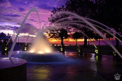 A fountain with illuminated water jets against a vibrant sunset sky, surrounded by trees and a waterfront view.