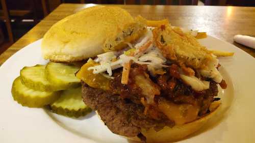 A hearty burger topped with coleslaw and barbecue sauce, served with pickles on the side.