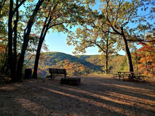 A serene outdoor scene with benches and a picnic table surrounded by colorful autumn trees and rolling hills.