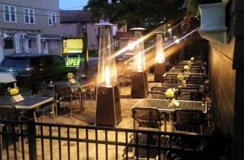 Outdoor dining area with tables, chairs, and patio heaters, illuminated at dusk, with a sign indicating it's open.