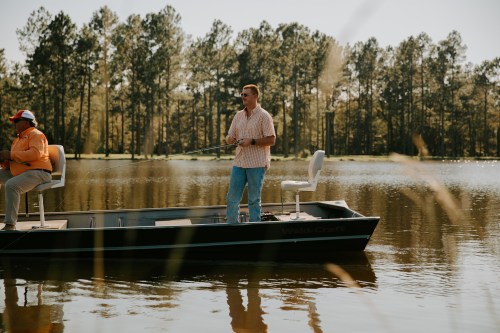 A man stands on a fishing boat, casting a line, while another person sits nearby on a calm lake surrounded by trees.