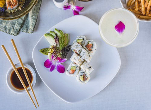 A plate of sushi rolls with avocado and salmon, garnished with an orchid, chopsticks, and a drink in the background.
