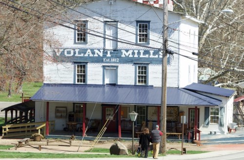 Historic Volant Mills building with a blue roof, surrounded by trees and power lines, featuring a porch and outdoor displays.
