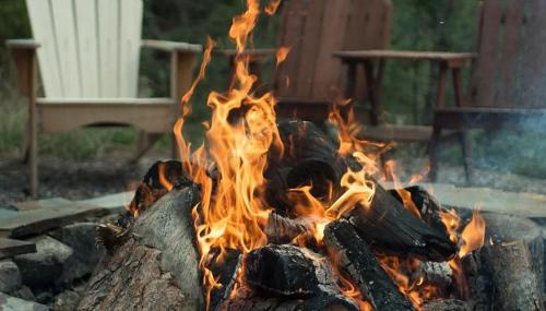A crackling campfire with bright flames, surrounded by wooden chairs in a natural outdoor setting.