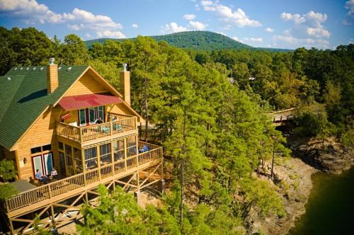 A wooden cabin surrounded by trees, overlooking a lake with a mountain in the background under a blue sky.