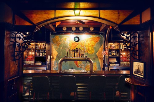 A cozy bar with a world map backdrop, wooden decor, and a row of beer taps in front of empty stools.