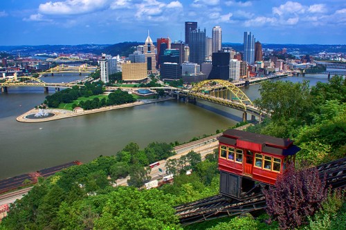 A red funicular descends a hill overlooking Pittsburgh's skyline and rivers, with bridges and greenery in the foreground.