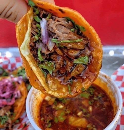 A hand holding a taco filled with meat, onions, and cilantro, next to a bowl of rich, spicy broth.