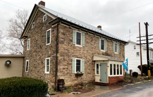 A two-story stone house with green shutters and a small porch, set against a cloudy sky.