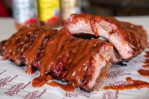 Close-up of juicy barbecue ribs drizzled with sauce, served on branded paper with bottles in the background.