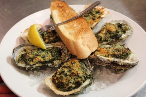 A plate of baked oysters topped with green herbs, served with a slice of toasted bread and a lemon wedge.