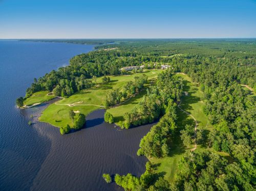 Aerial view of a lush green golf course bordered by a lake and dense forest under a clear blue sky.