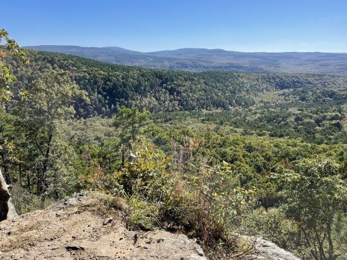 A scenic view from a rocky overlook, showcasing lush green hills and valleys under a clear blue sky.
