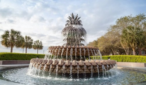 A decorative fountain shaped like a pineapple, surrounded by palm trees and lush greenery under a cloudy sky.