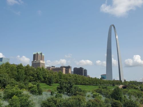 A tall arch monument stands against a blue sky, surrounded by greenery and city buildings in the background.