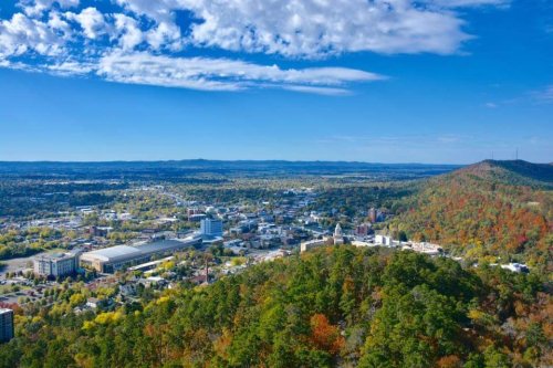 Aerial view of a vibrant city surrounded by green hills and colorful autumn foliage under a clear blue sky.