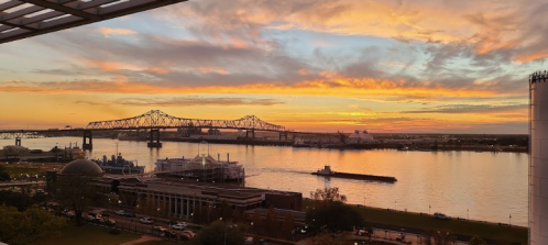 A vibrant sunset over a river, with a bridge in the background and a barge passing by.
