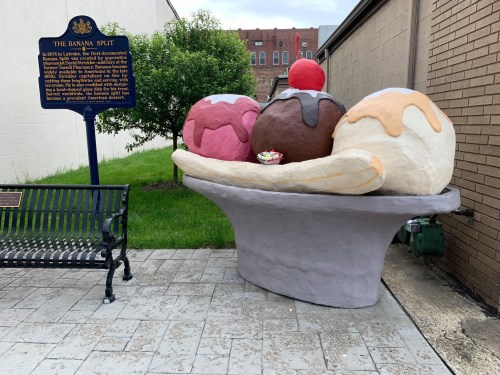 A large, colorful ice cream sundae sculpture sits on a bench, with a historical marker in the background.