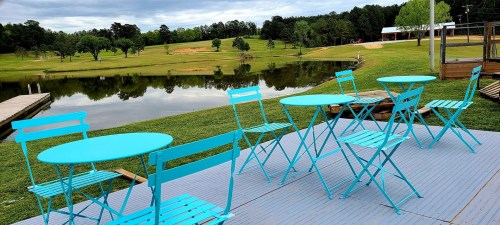 A serene outdoor scene featuring turquoise tables and chairs by a calm lake, surrounded by green grass and trees.