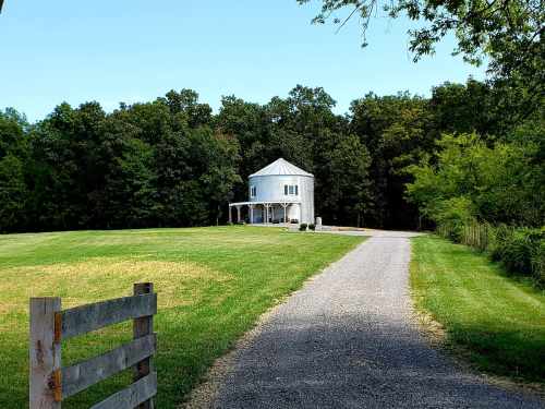 A white house with a porch sits at the end of a gravel driveway, surrounded by green grass and trees.