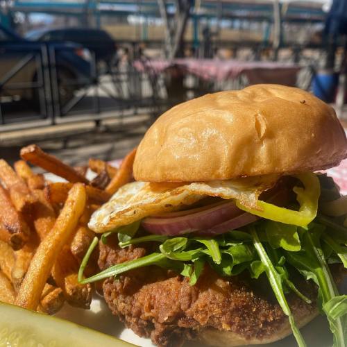 A crispy chicken sandwich with lettuce, tomato, and a fried egg, served with golden fries and a pickle on the side.