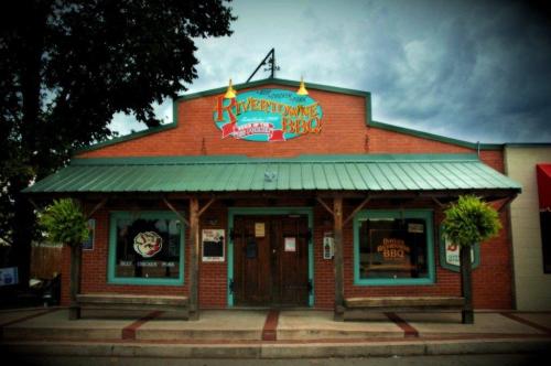 A brick building with a green roof, featuring a sign that reads "Rivertown BBQ" and decorative plants by the entrance.
