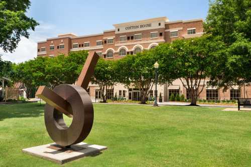 A modern sculpture in front of the Cotton House building, surrounded by green grass and trees on a sunny day.