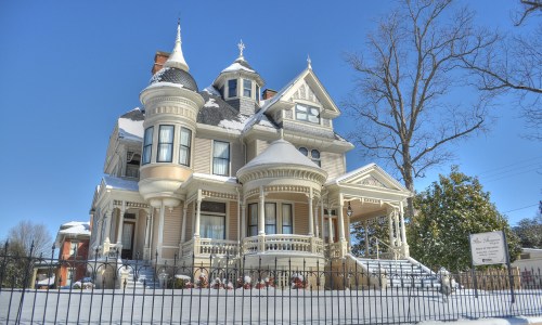 A large, ornate Victorian house covered in snow, featuring turrets and a wraparound porch against a clear blue sky.