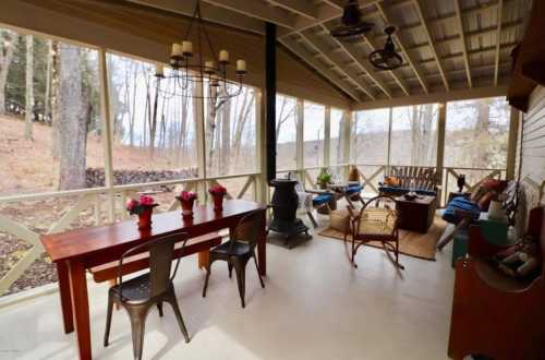 A cozy screened porch with a wooden table, chairs, and decorative plants, surrounded by trees in a serene setting.