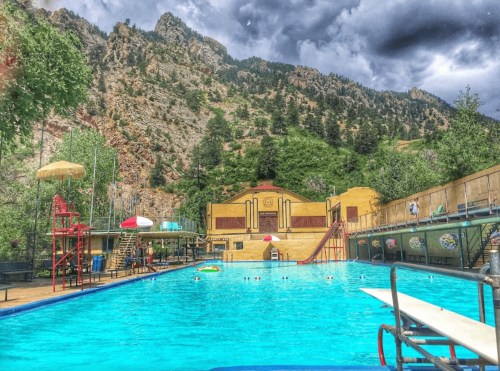 A vibrant outdoor swimming pool surrounded by mountains and trees, under a cloudy sky.