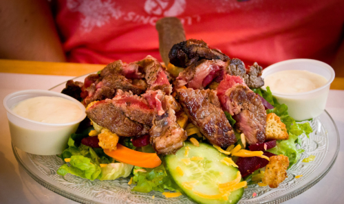 A plate of salad topped with grilled steak, cucumbers, cheese, and croutons, served with two small cups of dressing.