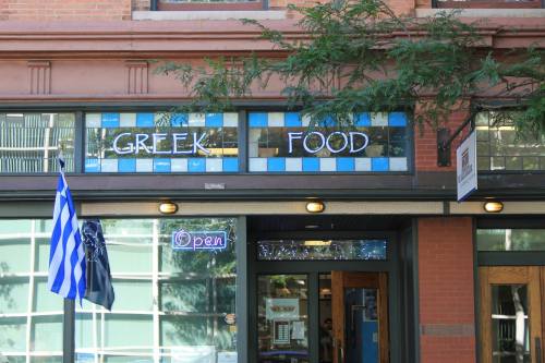 Exterior of a restaurant with "Greek Food" sign and an open neon sign, featuring a blue and white flag.