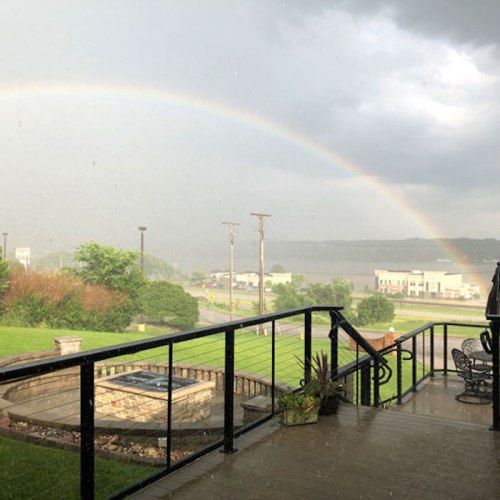 A rainbow arcs over a stormy landscape, viewed from a porch with a railing and potted plants. Rain falls softly.