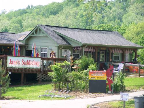 A quaint green building with a sign reading "Jane's Saddlebag," surrounded by greenery and outdoor seating.