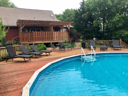 A wooden deck with lounge chairs surrounds a blue swimming pool, next to a house with a balcony and greenery.