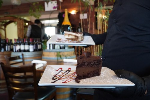 A server in black gloves holds two plates with chocolate cake and tiramisu, set in a cozy restaurant with wine bottles.