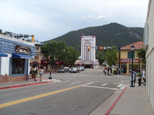 A quaint street scene with shops, people walking, and a mountain backdrop under a cloudy sky.