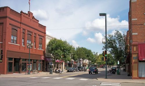 A street view of a small town with shops, trees, and vehicles under a partly cloudy sky.
