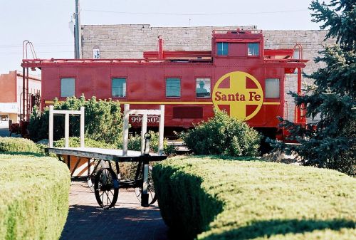 A red Santa Fe caboose displayed among green bushes and a wooden cart in a small town setting.