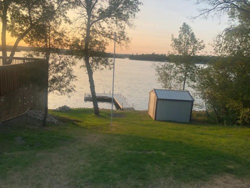 A serene lakeside view at sunset, featuring a dock, a small shed, and trees lining the shore.