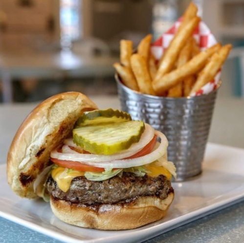 A juicy cheeseburger with pickles, lettuce, and tomato, served with a side of crispy French fries in a metal cup.