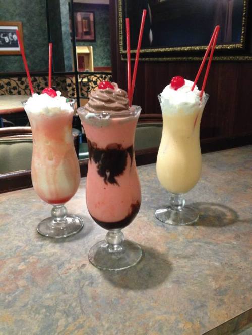 Three colorful milkshakes in tall glasses, topped with whipped cream and cherries, on a marble countertop.