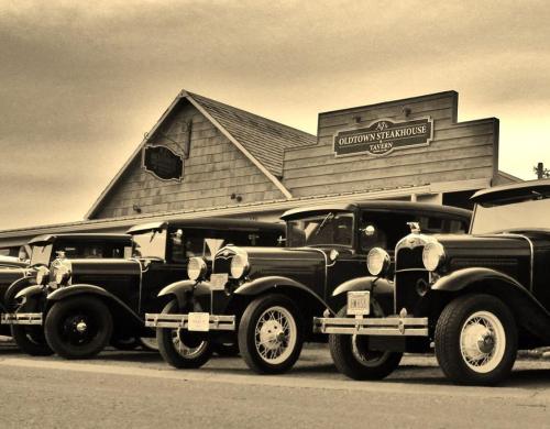 A row of vintage cars parked in front of Oldtown Steakhouse, with a cloudy sky overhead.