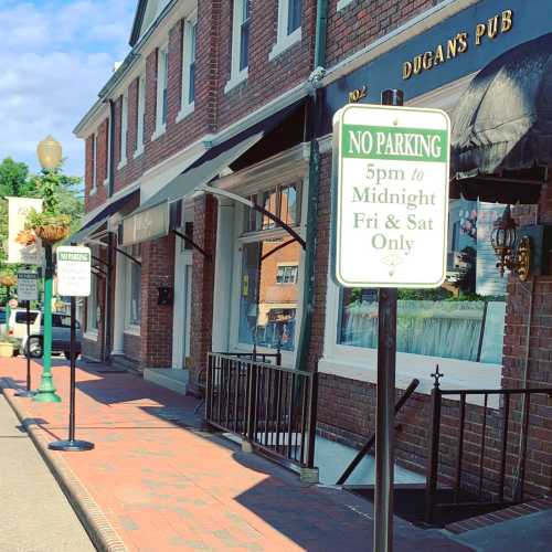 A street view of Dugan's Pub with a no parking sign and shops lining the sidewalk under a clear blue sky.