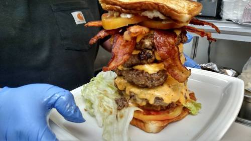 A towering burger stacked with multiple patties, bacon, cheese, lettuce, and tomato, served on a plate.