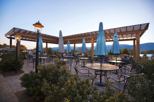Outdoor dining area with tables and blue umbrellas, surrounded by greenery and mountains at sunset.