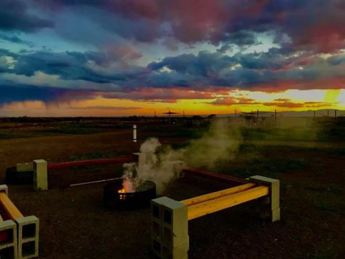 A cozy campfire surrounded by benches, with a colorful sunset and dramatic clouds in the background.