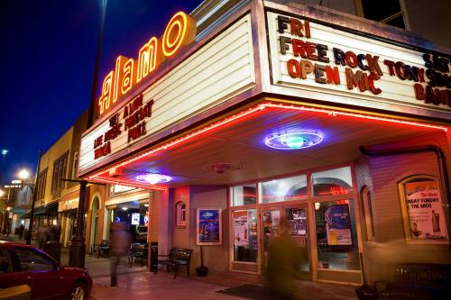 The Alamo theater marquee lit up at night, featuring events like open mic and free rock shows.