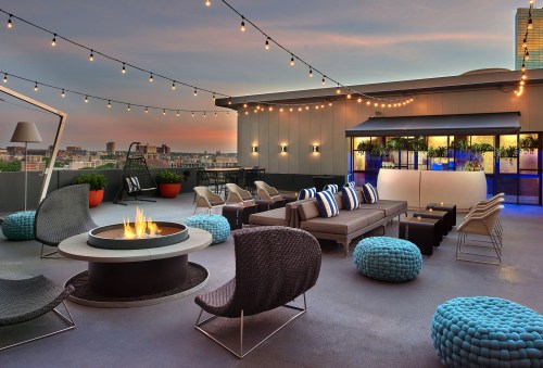 Rooftop lounge with cozy seating, a fire pit, and string lights, overlooking a city skyline at sunset.