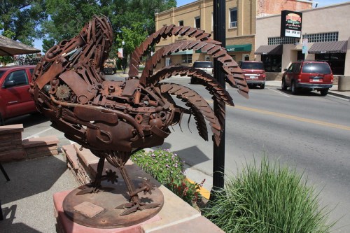 A large metal rooster sculpture made from various scrap materials, displayed on a sidewalk near a street.
